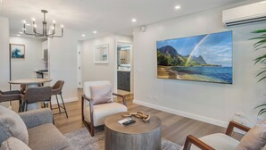 A modern living room with a dining area, a large painting of a tropical landscape, and a chandelier.