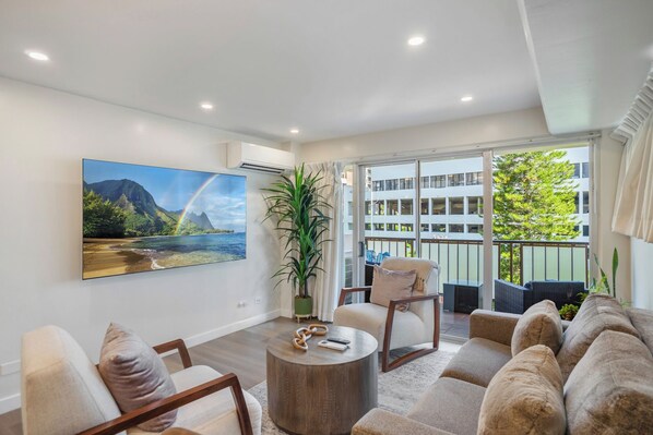 A spacious living room with a large window, a potted plant, and a painting of a beach scene.