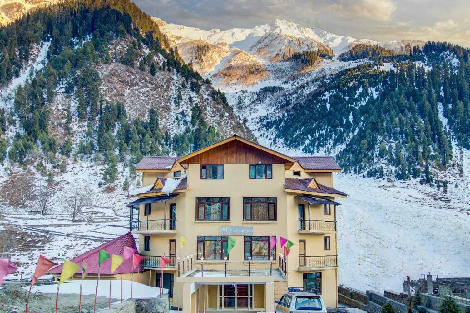 A multi-story hotel with balconies, set against a mountainous backdrop with snow-covered peaks.
