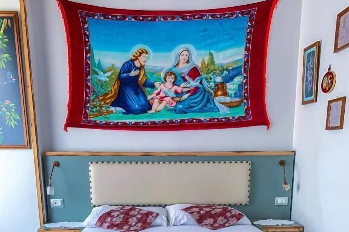 Sängyssä on valko-punainen kukallinen päiväpeite, sininen pääty ja sen yläpuolella uskonnollinen seinävaate.