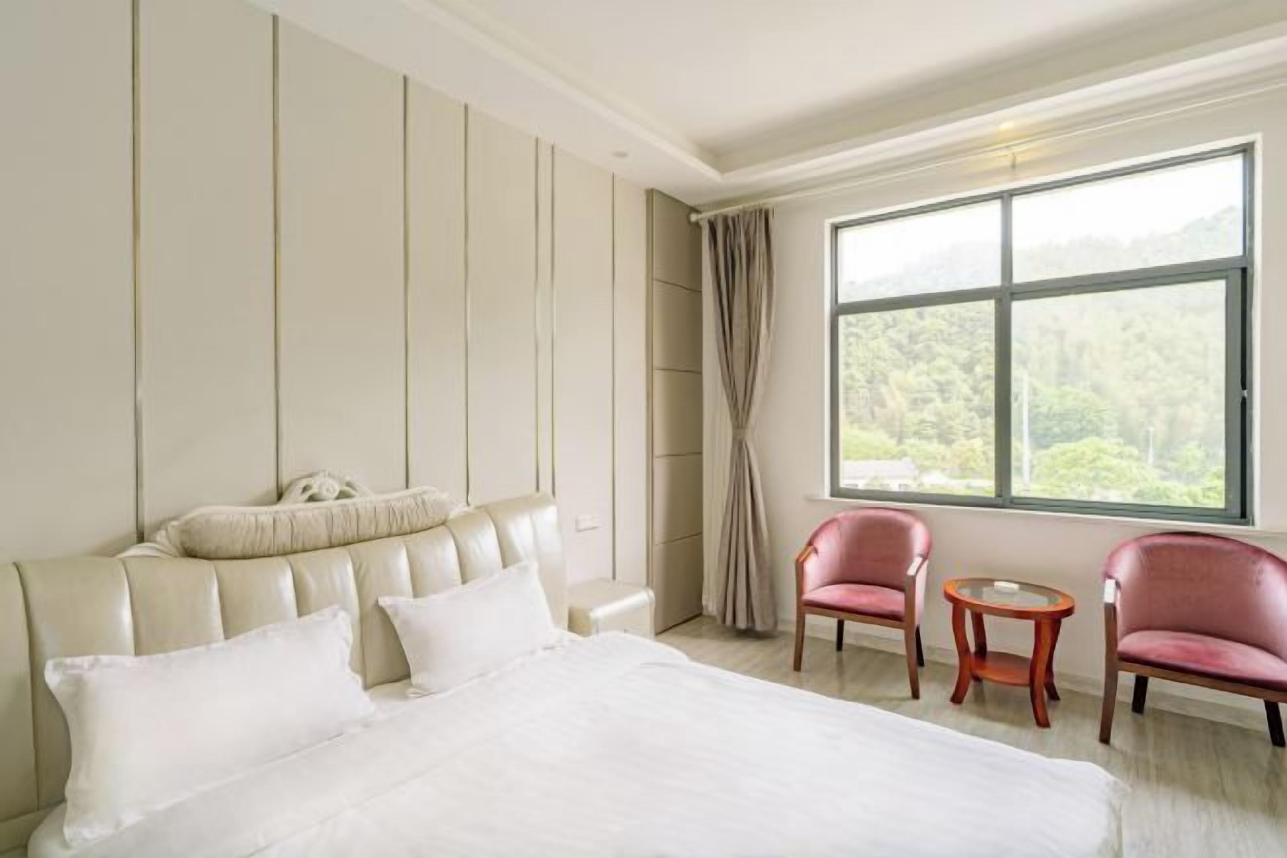 A hotel room with a large bed, two pink armchairs, a small round table, and a large window with a view of greenery.
