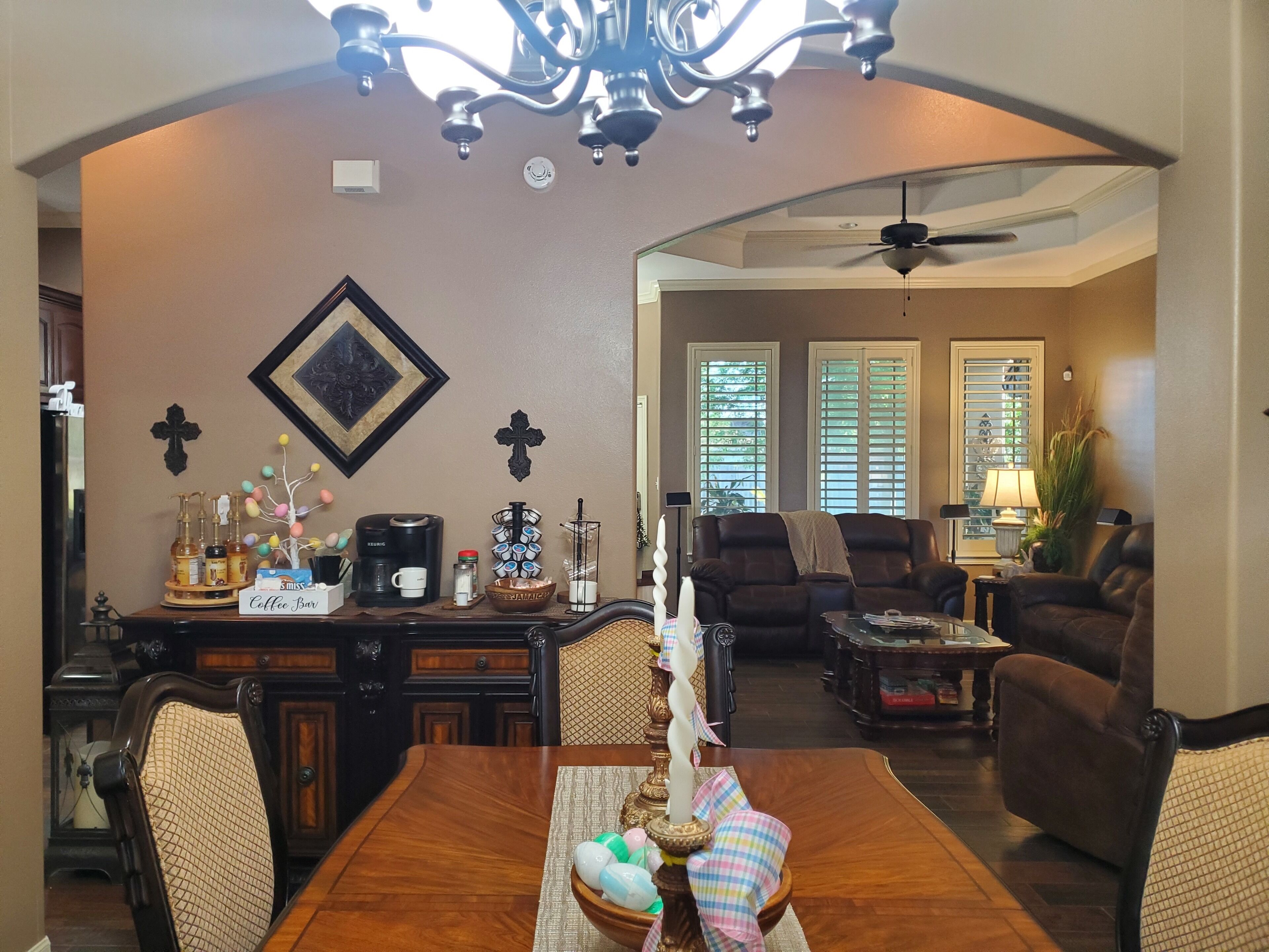 A dining area with a wooden table, chairs, and a chandelier. There is a coffee station with a coffee maker and cups. The room has a cross on the wall and a window with blinds.