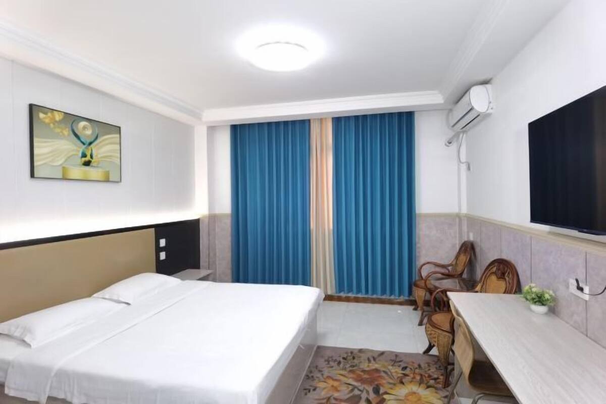 Quarto de hotel com cama, escrivaninha, cadeira, janela com cortinas e ar condicionado fixado na parede.