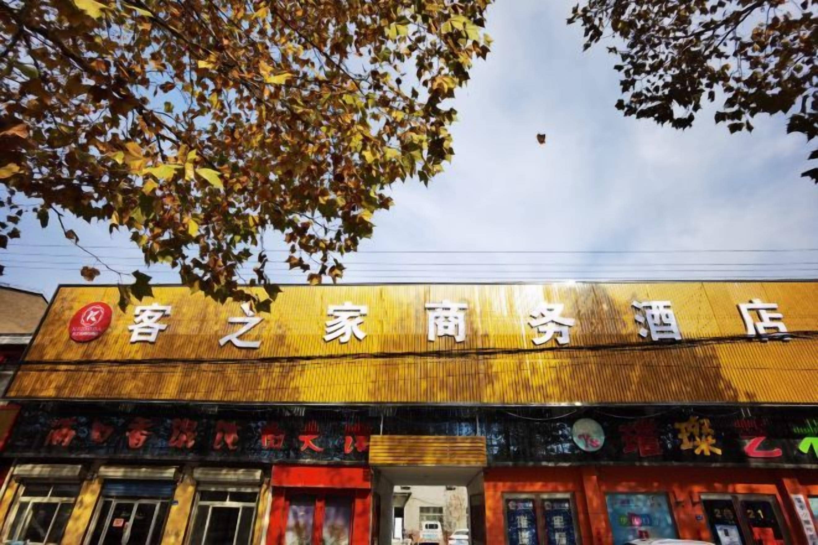 Ein Hotel mit einem Schild in chinesischen Schriftzeichen, eine Ladenzeile mit verschiedenen Geschäften und ein Baum mit Herbstlaub.