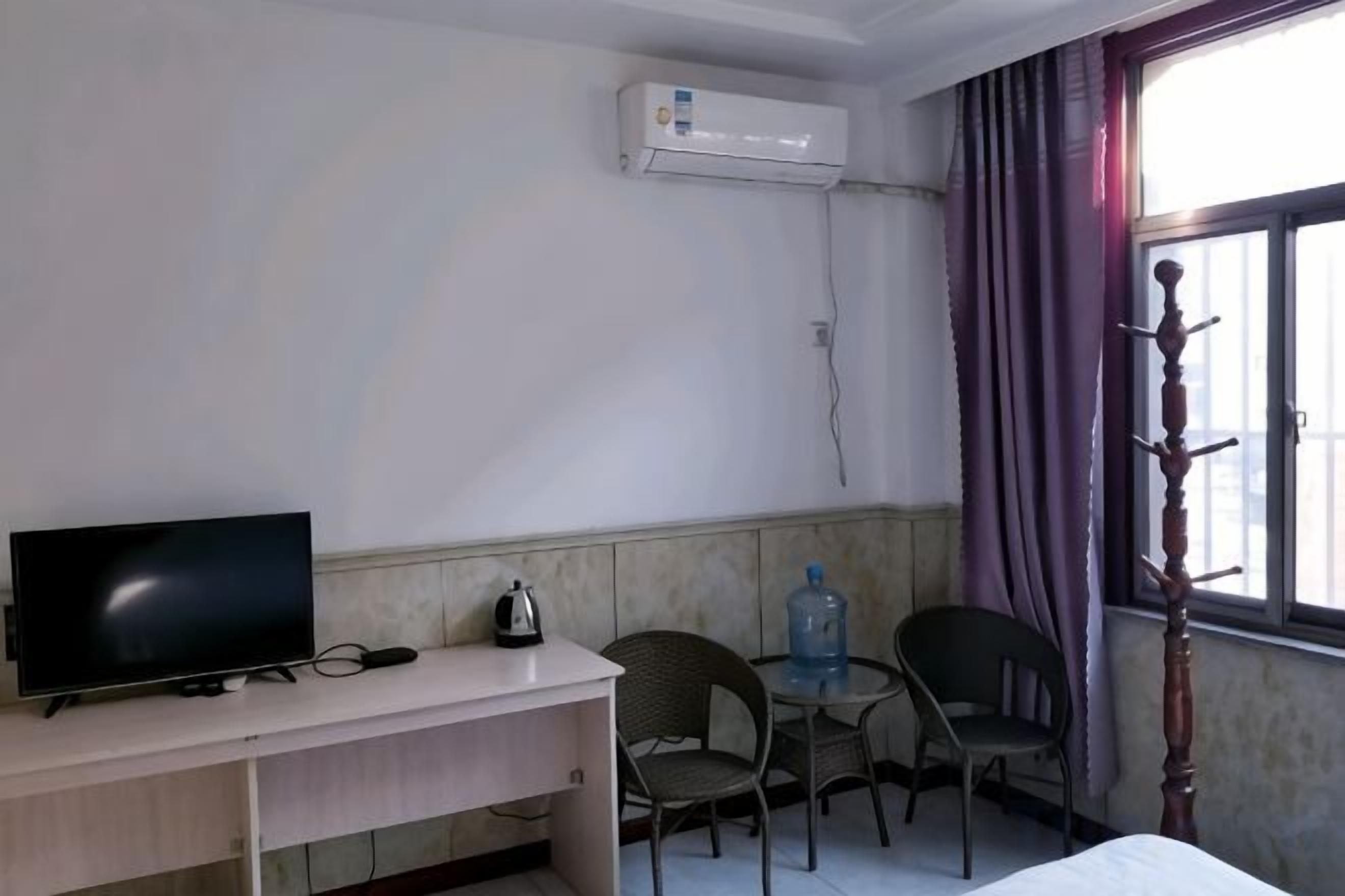 Una habitación con cama, escritorio con televisor, dos sillas, equipo de aire acondicionado y ventana con cortinas.