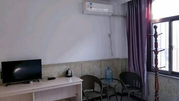 Una habitación con cama, escritorio con televisor, dos sillas, equipo de aire acondicionado y ventana con cortinas.