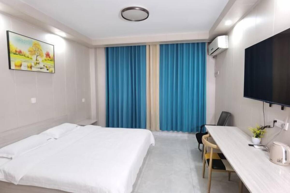 Quarto de hotel com cama, escrivaninha, cadeira, televisão e janela com cortinas.