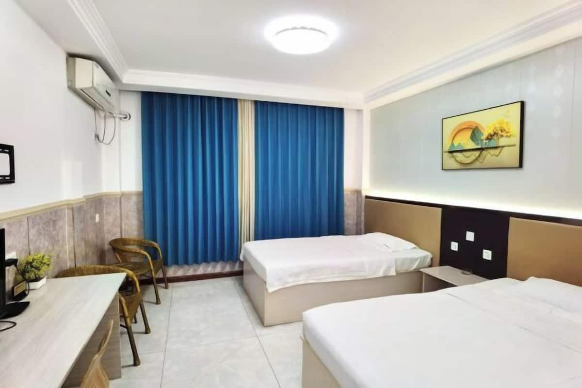 Quarto de hotel com duas camas, uma escrivaninha, uma cadeira e uma janela grande com cortinas azuis.