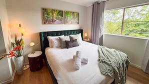 A bedroom with a large bed, a ceiling fan, two wall-mounted paintings, a bedside table with a lamp, and a window with a view of trees. - Leaf Luxe - 9 Pools BBQ Gym (Cairns)