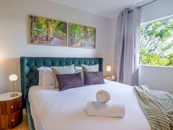 A neatly made bed with a green upholstered headboard, white linens, and a rolled towel. There are two bedside tables with lamps, and two framed pictures of a forest on the wall.