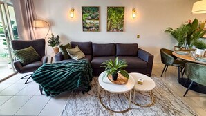 A modern living room with a dark sofa, two side tables, and a coffee table. There are two wall-mounted light fixtures and two framed pictures on the wall. - Leaf Luxe - 9 Pools BBQ Gym (Cairns)