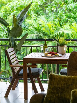 A wooden table with a bottle, glass, and food items on a balcony with a view of lush greenery. - Leaf Luxe - 9 Pools BBQ Gym (Cairns)