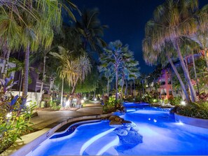 A nighttime resort pool area with palm trees and a water feature.