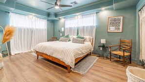 Un dormitorio con una cama de madera, dos ventanas con cortinas blancas, un ventilador de techo y una mesita de noche pequeña con una lámpara.