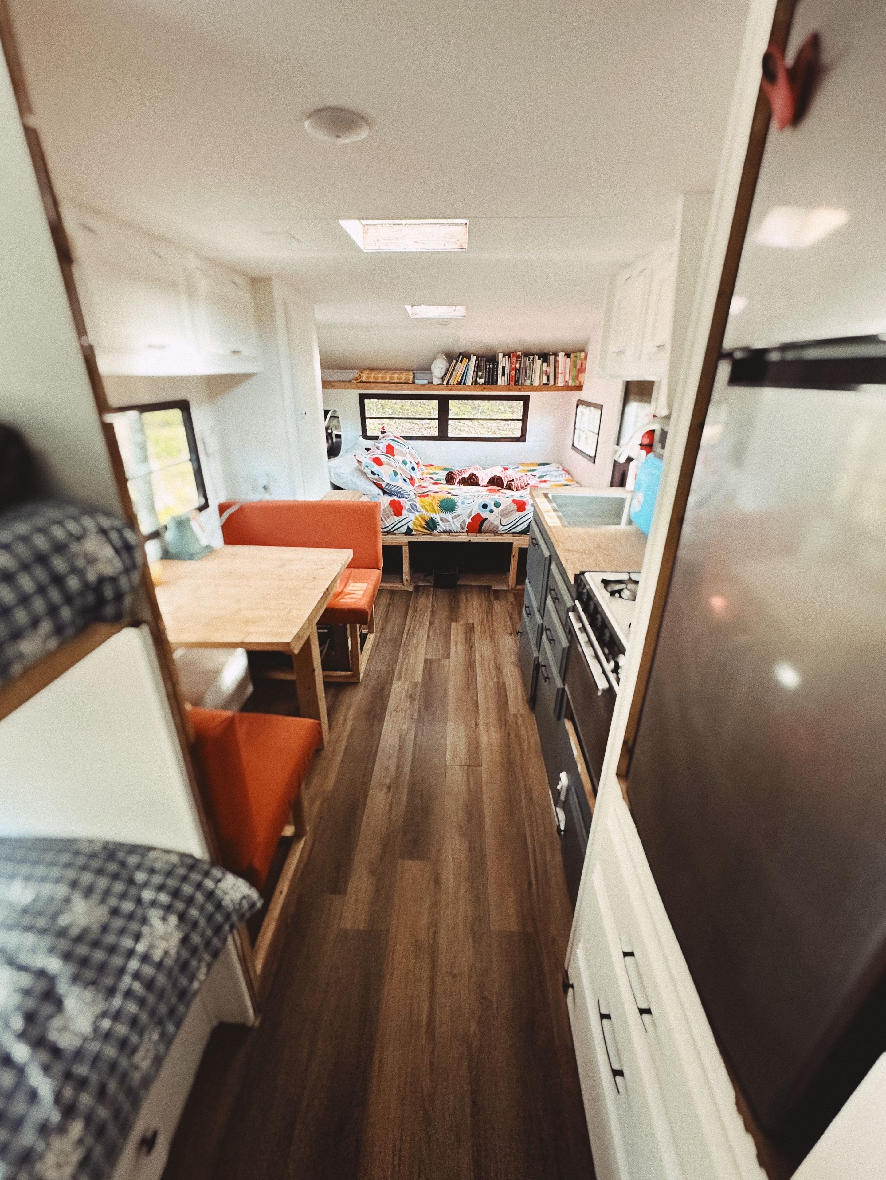 A compact RV interior with a kitchenette, a bed with colorful bedding, a dining area, and a small seating space.