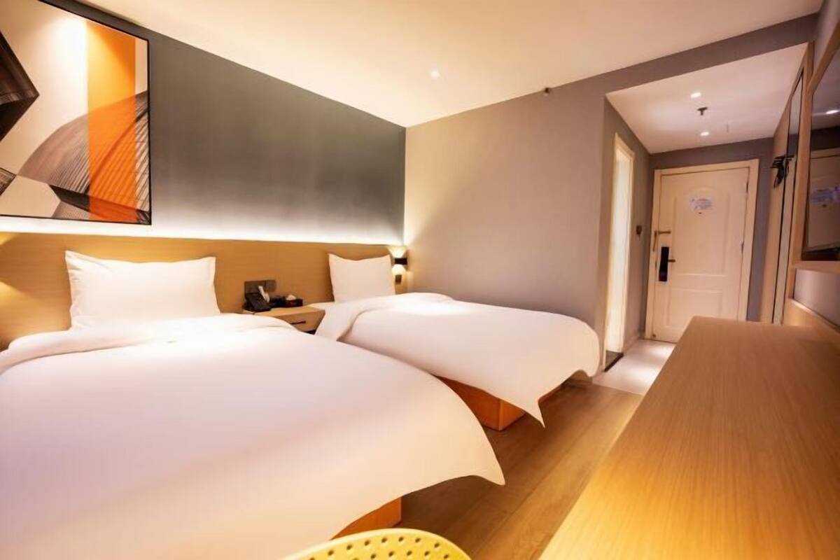 Habitación de hotel con dos camas, un escritorio de madera, una mesita de noche y una lámpara de pared.