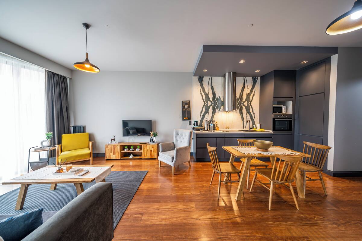 Una sala de estar moderna con sofá, mesa de comedor y sillas, un área de cocina con electrodomésticos, y un ventanal con cortinas.