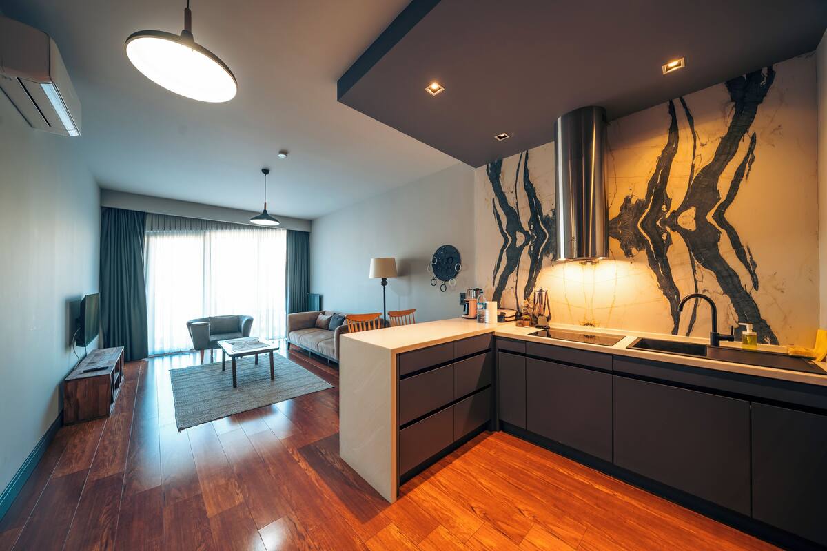 Una cocina moderna con una isla central, un comedor con sofá y sillas, y una sala de estar con un ventanal y cortinas.