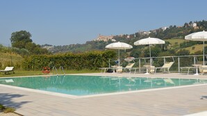 Piscina com espreguiçadeiras e guarda-sóis, cercada por vegetação e com vista para uma encosta com edifícios.