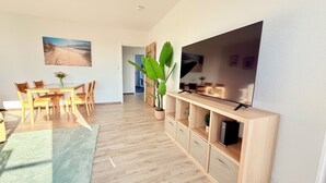 A modern living room with a wooden dining table, chairs, a large flat-screen TV mounted on a wooden console, and a large green plant.