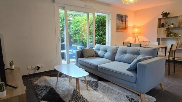 Ein modernes Wohnzimmer mit einem grauen Sofa, einem kleinen weißen Couchtisch und einem schwarz-weißen Teppich. Links ist ein Fernseher, rechts eine Lampe und im Hintergrund ein Essbereich zu sehen.