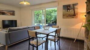 Ein modernes Wohnzimmer mit einer Couch, einem Esstisch, Stühlen, einem Fernseher und großen Fenstern mit Blick auf Grünflächen.