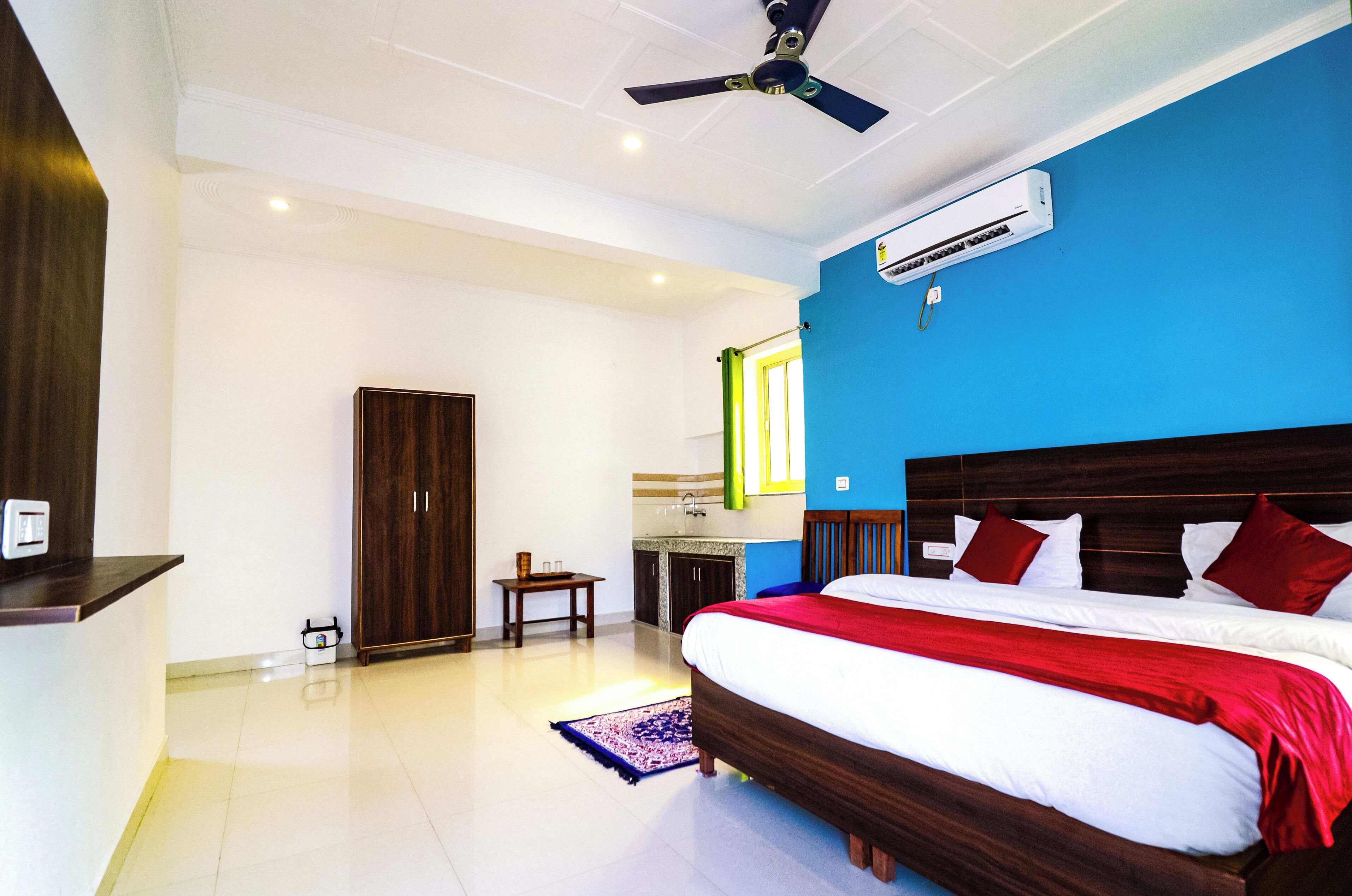 A bedroom with a large bed, a ceiling fan, an air conditioning unit, a wooden wardrobe, a small table, and a bathroom visible through an open door.
