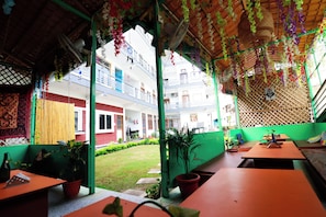 An outdoor seating area with tables and chairs, potted plants, and a view of a building with balconies. - Manzil Hostel (Rishikesh)