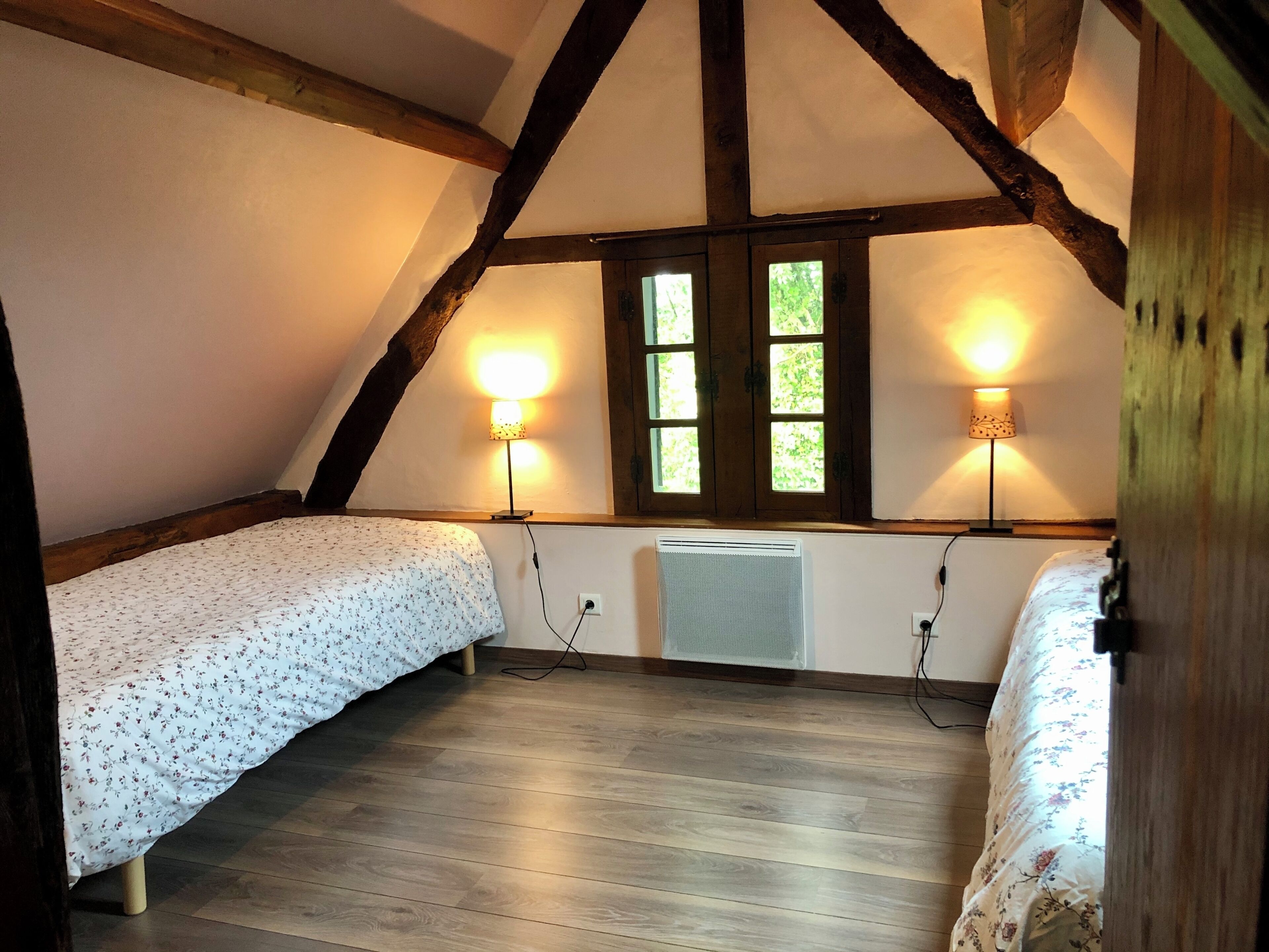 A bedroom with two beds, wooden beams, a sloped ceiling, and a window with a view of greenery.