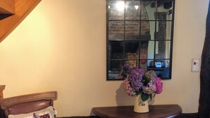 A cozy corner with a wooden chair, a table with a vase of flowers, a stone wall, and a large arched window reflecting the interior. - Renovated bread oven in pretty Normandy property (Étréville)