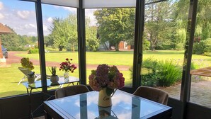 A sunlit room with a glass table and chairs, a vase of flowers, and a view of a green garden and trees. - Renovated bread oven in pretty Normandy property (Étréville)