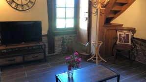 A living room with a large wall clock, a television, a vase with flowers, a staircase, and a chair with a cushion. - Renovated bread oven in pretty Normandy property (Étréville)
