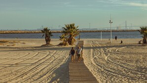 Un paseo marítimo de madera que lleva a una playa con palmeras y un puente distante.
