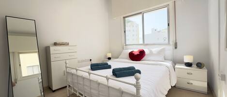 A bedroom with a white bed, a dresser, a nightstand, a mirror, and a window with a view of buildings.