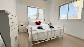 Ein Schlafzimmer mit einem weißen Bett, zwei Nachttischlampen, einem roten herzförmigen Kopfkissen und Blick auf Gebäude durch große Fenster.