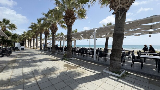Eine Terrasse am Meer mit Tischen und Stühlen unter weißen Sonnenschirmen, Palmen und Blick aufs Meer.