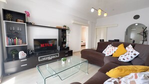 Ein modernes Wohnzimmer mit einem Flachbildfernseher, der "NETFLIX" anzeigt, einem Glastisch, einem braunen Sofa mit gemusterten Kissen und einem Bücherregal mit Dekorationselementen.