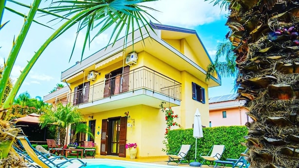 Una casa gialla a due piani con un balcone, una piscina e poltrona relax.