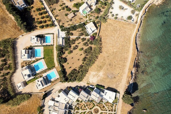 An aerial view of a coastal property with multiple modern houses, a large sandy area, and a clear blue sea. - Twin Suns (Drios)