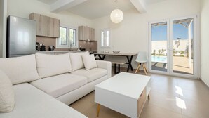 Un saló modern amb un sofà, una taula de centre i una televisió. Hi ha una zona de cuina amb armaris i una taula de menjador. L'espai disposa de grans finestres i vistes a l'exterior.
