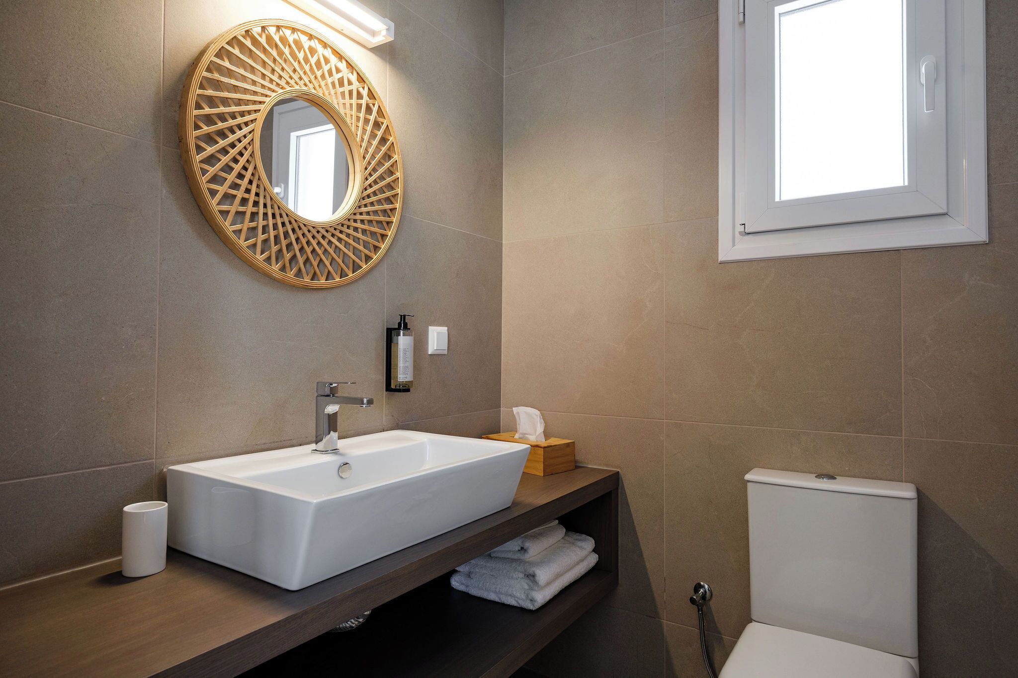 Een moderne badkamer met een rechthoekig wastafelmeubel, een ronde spiegel met geometrische omlijsting, een toilet en een raam dat natuurlijk licht binnenlaat.