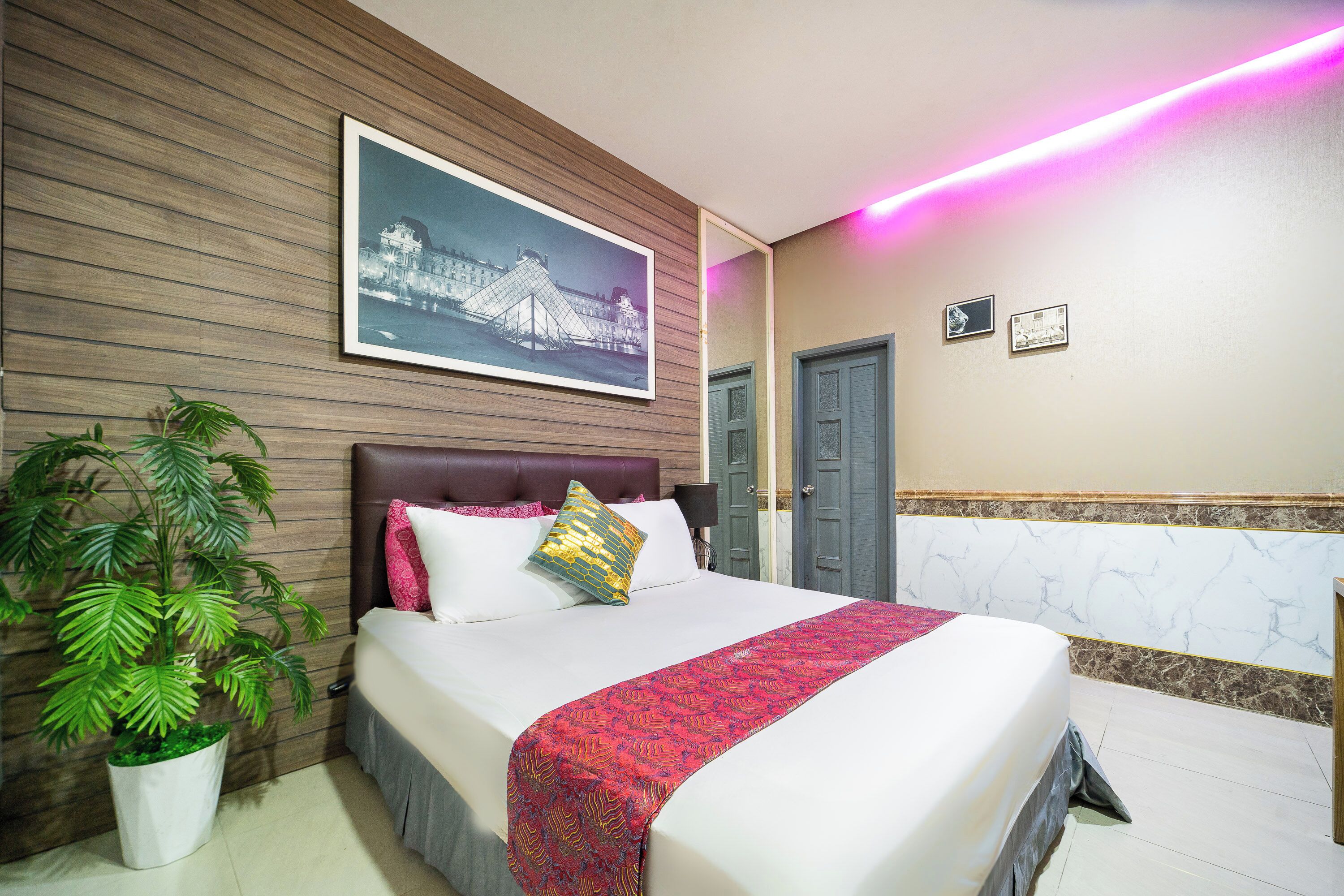 A hotel room with a bed, a large potted plant, a framed picture on the wall, and a marble accent wall.