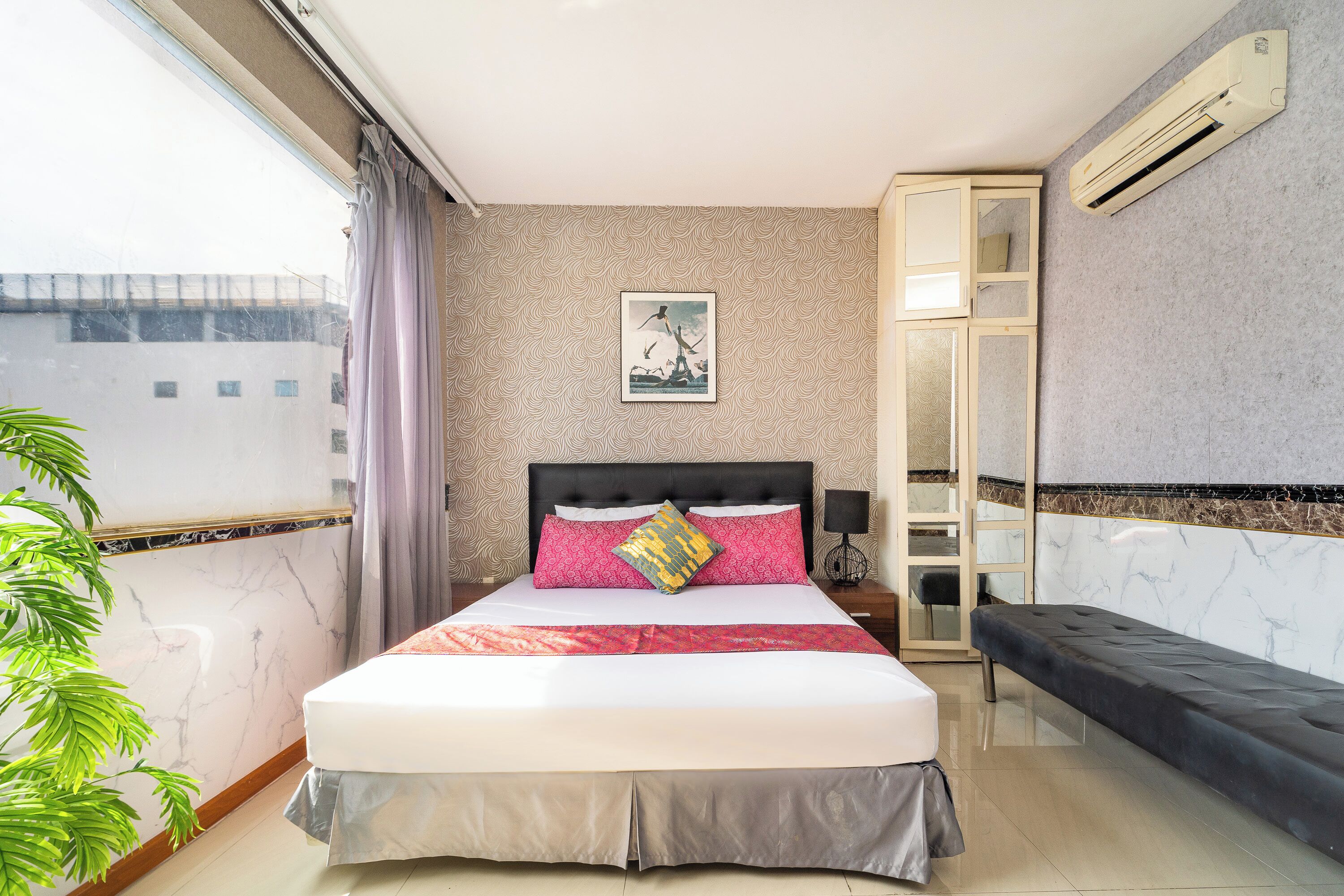 A modern hotel room with a large bed, a bench, a potted plant, and a view of the cityscape.
