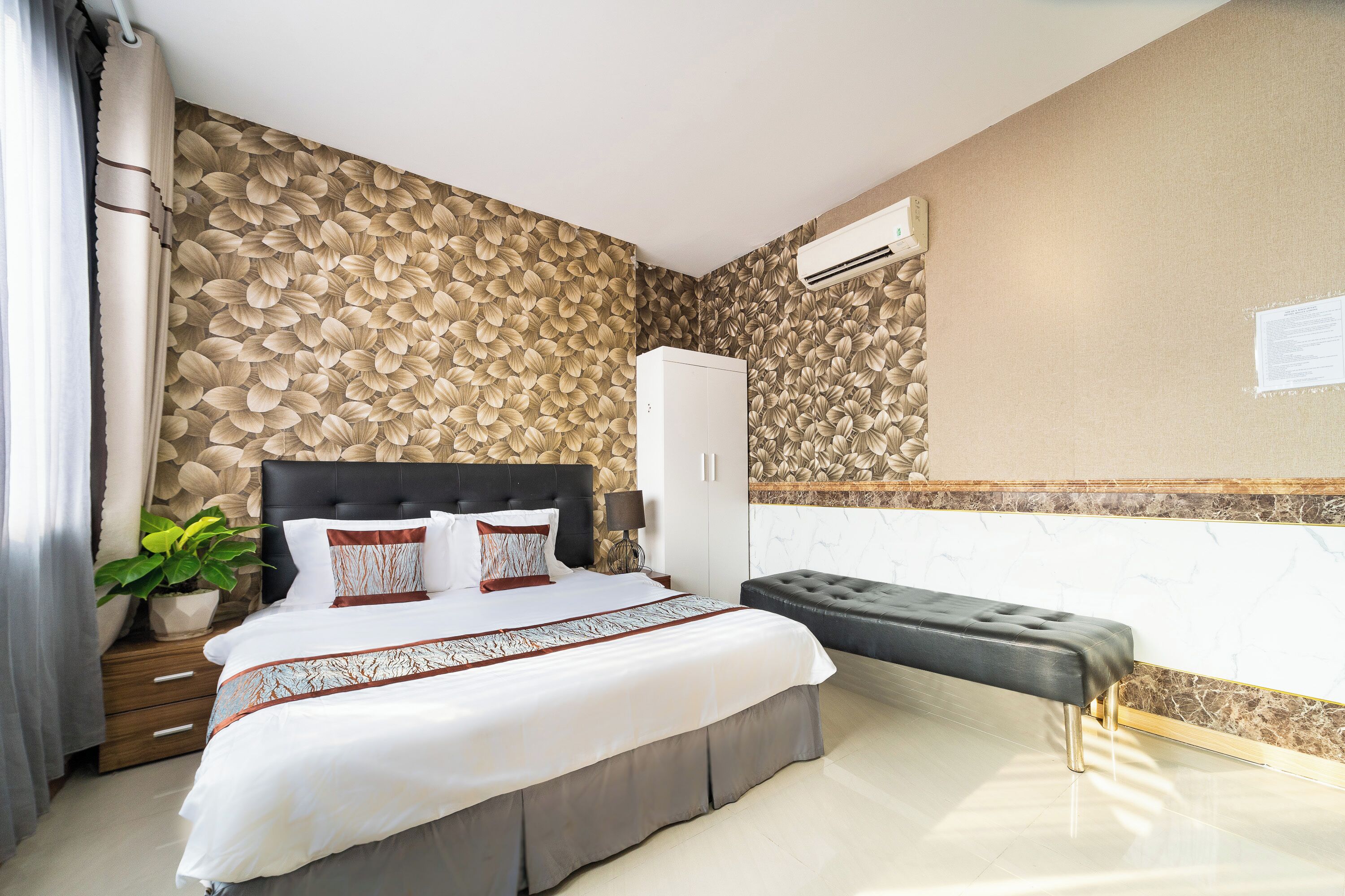A bedroom with a bed, a nightstand, a potted plant, a bench, and a wall-mounted air conditioner.