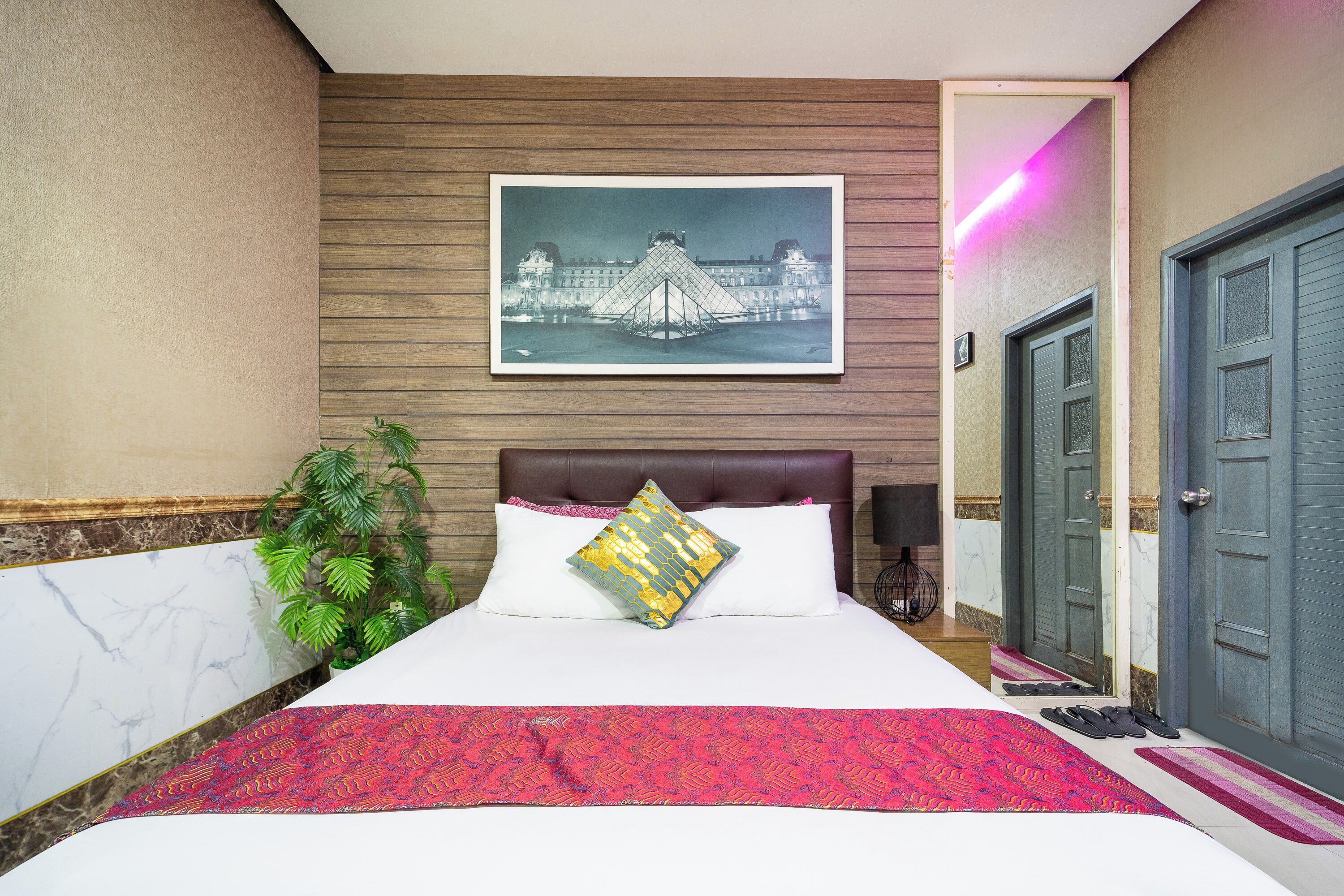 A hotel room with a bed, a large framed picture of a bridge, a potted plant, and a door.