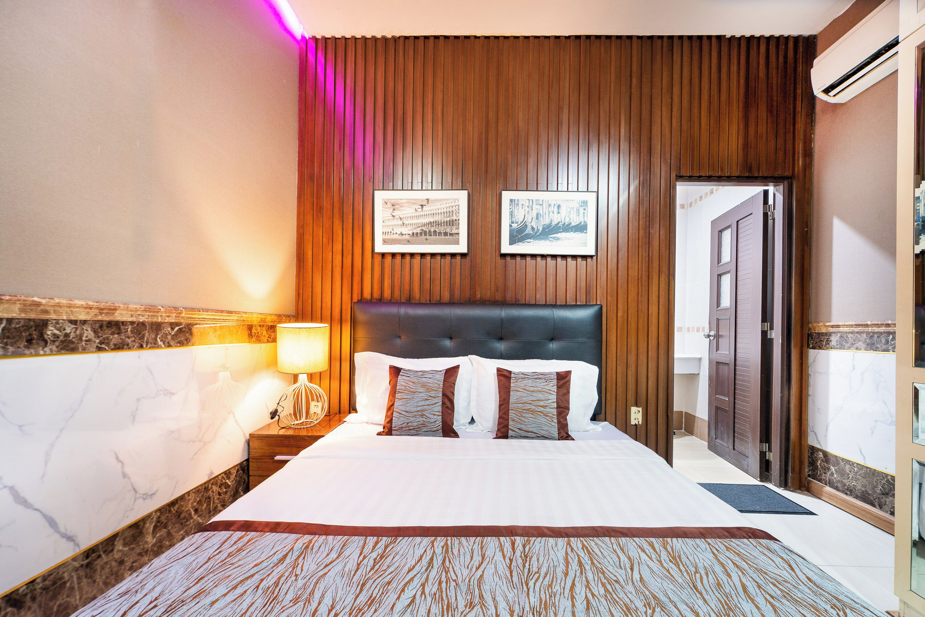 A hotel room with a large bed, wooden panel wall, two framed pictures, a nightstand with a lamp, and a door.