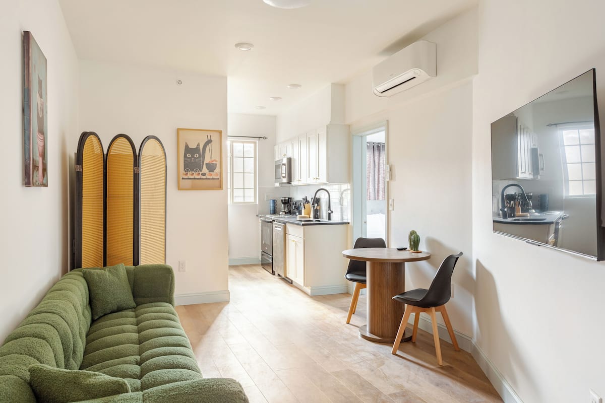 A modern living room with a green sofa, a dining area with a small table and chairs, a kitchen in the background, and a large mirror on the wall.
