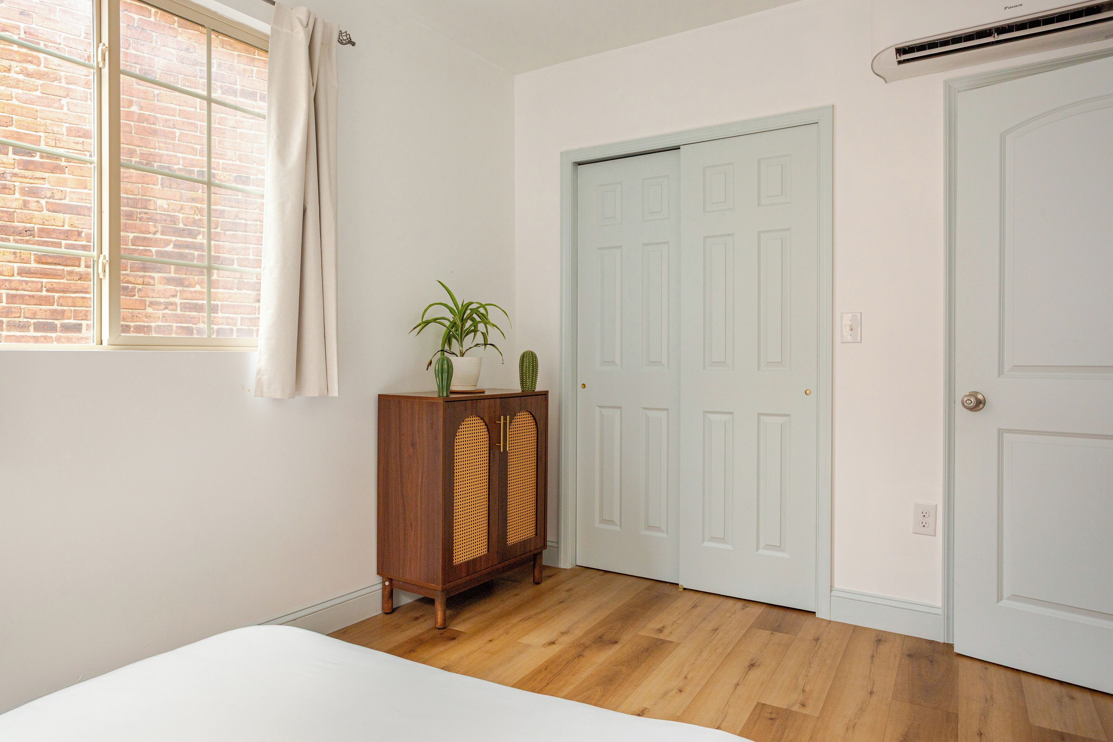 A room with a wooden floor, a white bed, a wooden cabinet with plants, and a white door with a window above it.