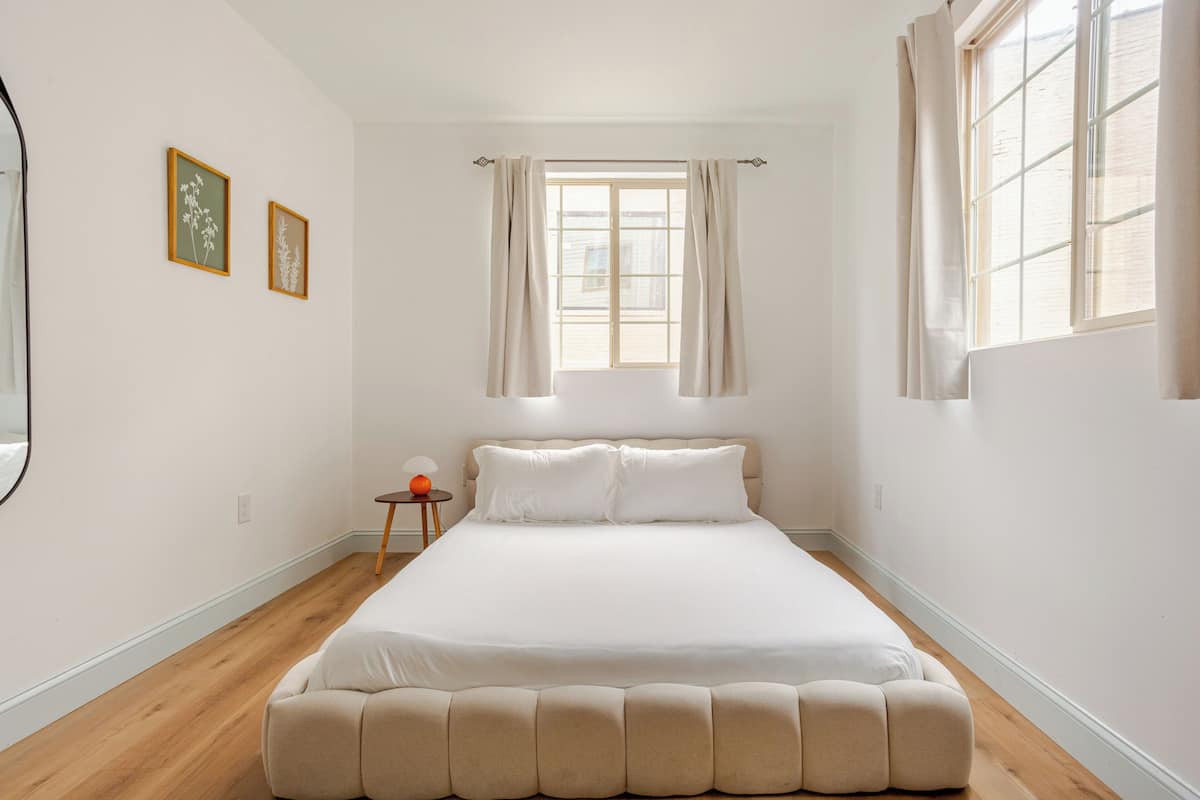 A bedroom with a large bed, a window with curtains, a small table with a lamp, and two framed pictures on the wall.