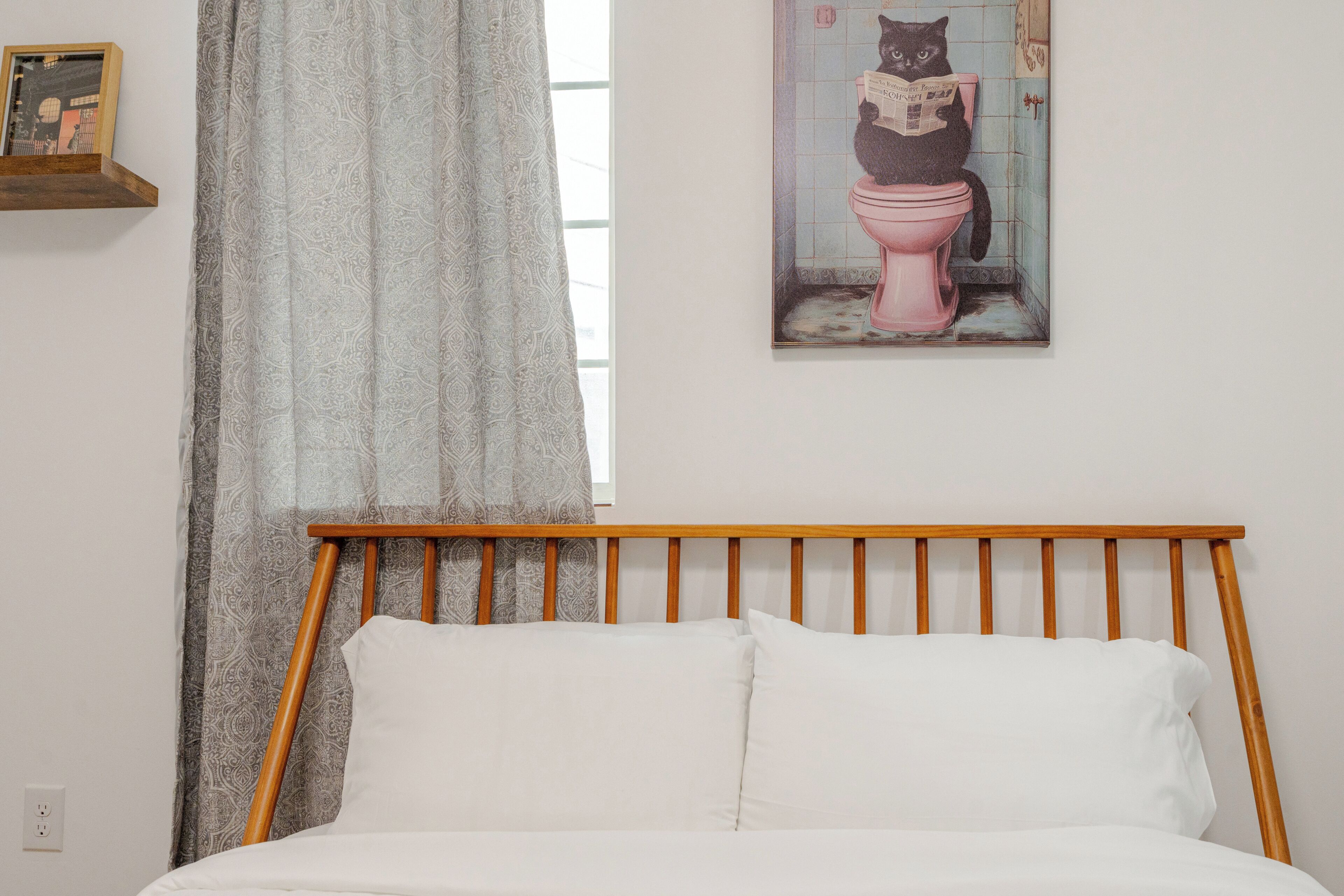 A bedroom with a wooden headboard, a bed with white bedding, a patterned curtain, and a wall-mounted picture of a cat reading a newspaper on a toilet.